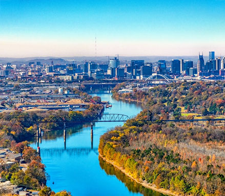 Aerial view of a river with bridges, surrounded by fall trees and a city skyline in the distance.